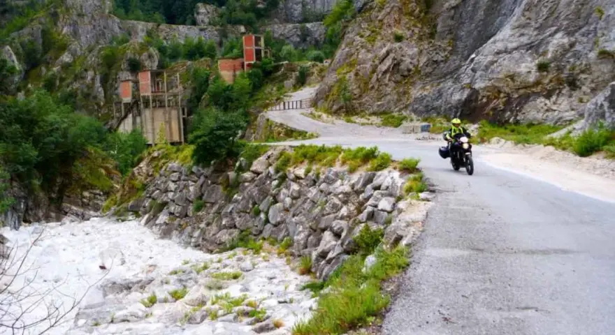 Un motociclista percorre una strada tortuosa tra le Alpi Apuane, con un fiume di sassi e ruderi industriali sullo sfondo.