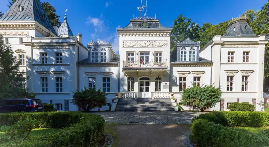 Elegancki pałac w Nosowie z wieżyczkami i ozdobnym balkonem, otoczony zielenią.