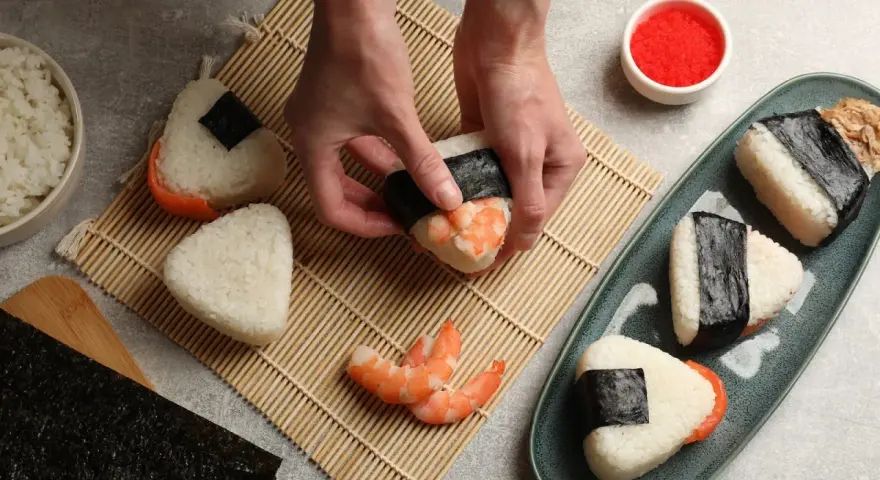 Ręce formują onigiri z krewetkami, tworząc pyszne japońskie jedzenie. Obok miska ryżu, nori i talerz gotowych przysmaków.