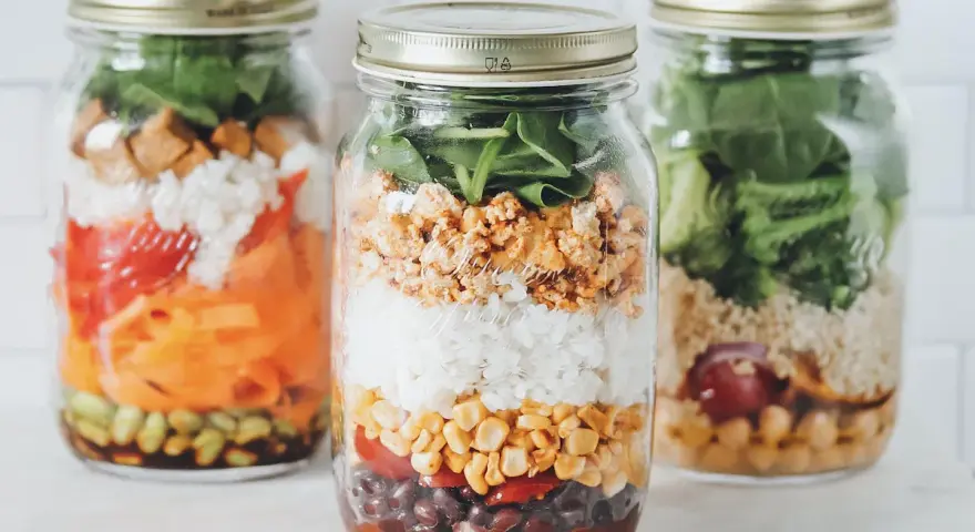 Drei bunte Schichtsalate im Glas, bereit für unterwegs. Jedes Glas ist ein kleines Kunstwerk mit Reis, Gemüse und Proteinen.