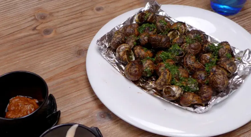 Caracoles con mantequilla y perejil, servidos en papel de aluminio sobre un plato blanco, junto a un cuenco con salsa.