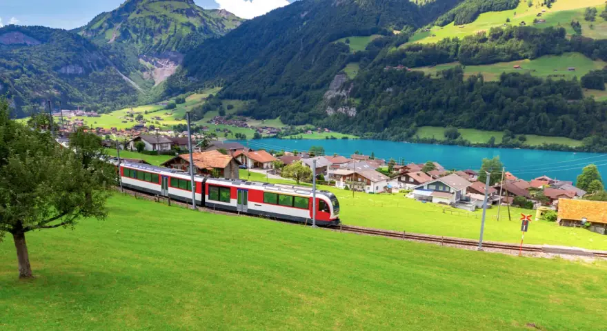 A red and white train travels along the **Golden Pass Line Lucerne to Interlaken**, passing a village by a turquoise lake, with lush green mountains in the background.