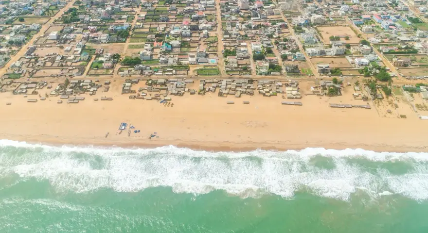 Widok z lotu ptaka na plażę i zabudowania w Kotonu, stolicy Beninu. Zielone morze z falami obmywa piaszczysty brzeg, za którym rozciąga się miasto.
