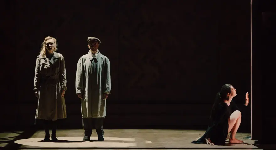 Magdalena Twardowska w scenie teatralnej. Dwie postacie w płaszczach stoją w świetle, obok nich kobieta w czarnej sukni klęczy przy pionowym świetle.