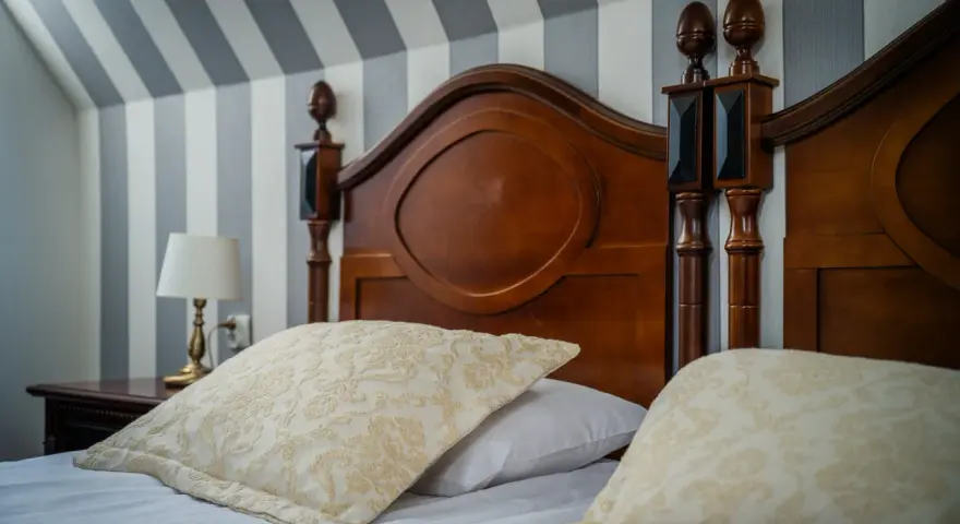 Eleganckie łóżko z drewnianym zagłówkiem i ozdobnymi poduszkami. Ciekawe, ile wynosi ryczałt za nocleg w takim miejscu?