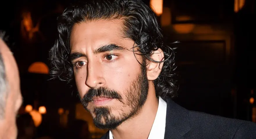 Primer plano de un hombre con cabello oscuro y rizado, y una barba poco poblada larga. Viste una camisa blanca y un traje oscuro.