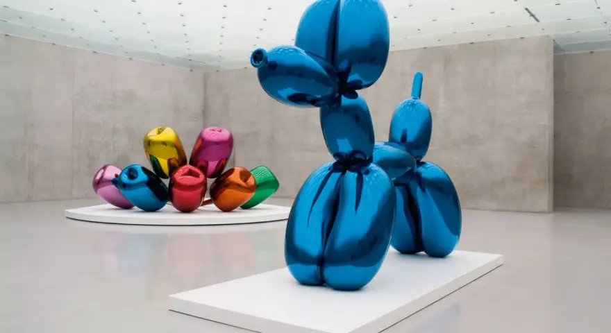 Un chien ballon bleu iconique de Jeff Koons, reflétant l'espace d'exposition. D'autres sculptures de ballons colorés sont visibles en arrière-plan.
