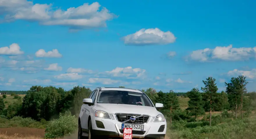 Białe Volvo XC60 T6 jedzie po piaszczystej drodze wśród zieleni, pod błękitnym niebem z chmurami.