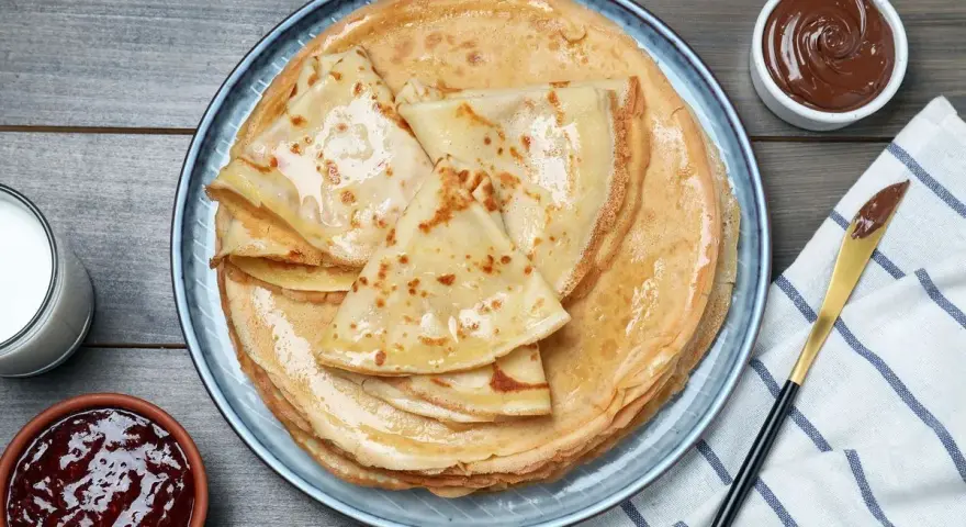 Pile de crêpes dorées, parfaites pour une recette pâte à crêpes. Accompagnées de confiture, pâte à tartiner et verre de lait.