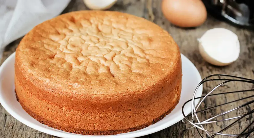 Perfekter Biskuitboden: Ein goldbrauner Kuchen, bereit für Ihr nächstes biskuitboden rezept. Eier und Schneebesen deuten auf die Zubereitung hin.