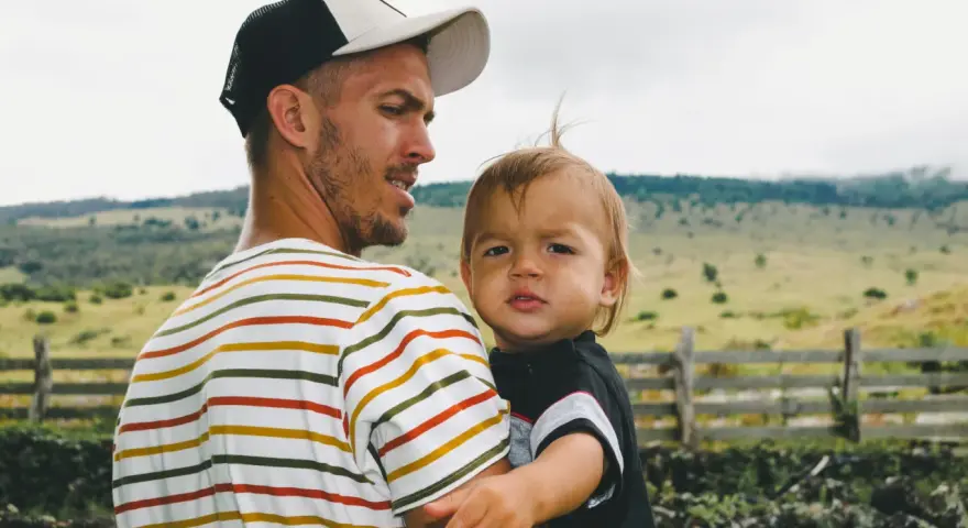 Ein Mann mit Bart und Kappe trägt ein Kind. Die Eigenschaften eines Papas zeigen sich in seiner liebevollen Umarmung.