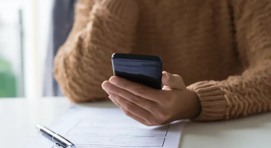 Persona revisa su teléfono móvil, modelo 035, mientras tiene un bolígrafo y un formulario de solicitud sobre la mesa.