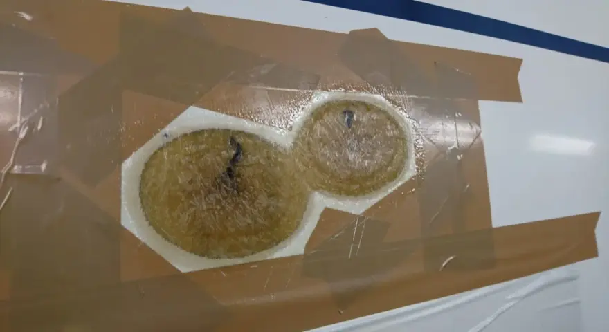 Reparaturstelle an einem Boot, bereit für das gelcoat erneuern. Zwei runde, faserige Füllungen sind mit Klebeband abgedeckt.