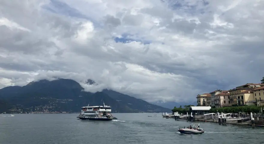 Promenada nad jeziorem Como, z górami w tle i statkiem wycieczkowym. W oddali widać urokliwe miasteczko.