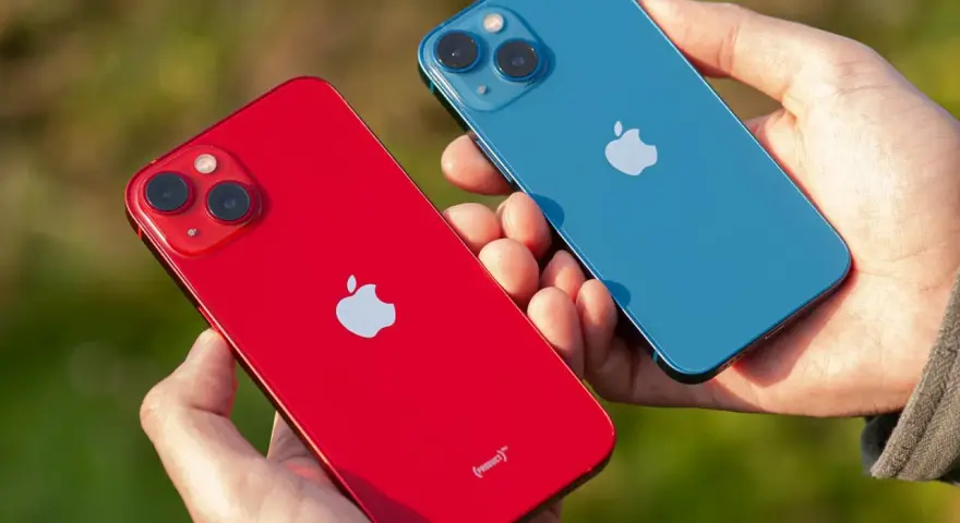 Dwie dłonie trzymają dwa telefony: czerwony i niebieski iPhone 13 mini. Aparat obu modeli jest widoczny.