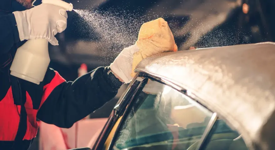 A person in gloves sprays a convertible car's fabric top with cleaning solution, then wipes it with a sponge. This is how you wash a convertible car.
