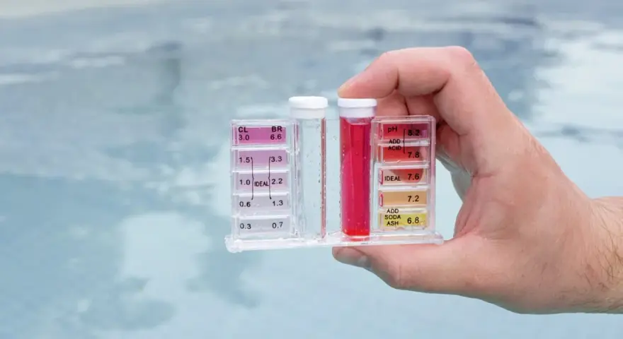 Hand holds pool water test kit. Red liquid in the pH tube indicates a high reading, suggesting acid is needed to lower pH.