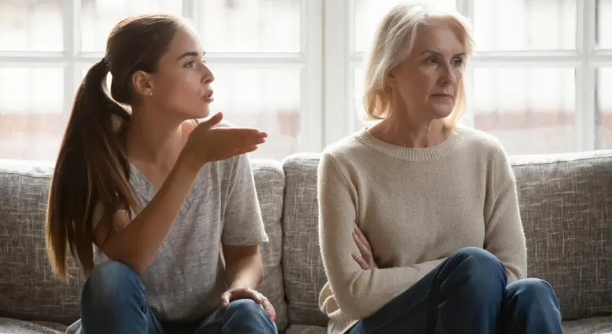 Junge Frau gestikuliert wütend, während ihre Mutter abgewandt sitzt. Es scheint, als ob meine Tochter mich hasst.