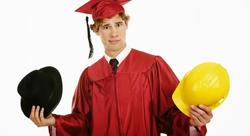 Studente universitario lavoratore con toga e cappello da laureato, tiene in mano un cappello nero e un casco giallo, indeciso sul futuro.