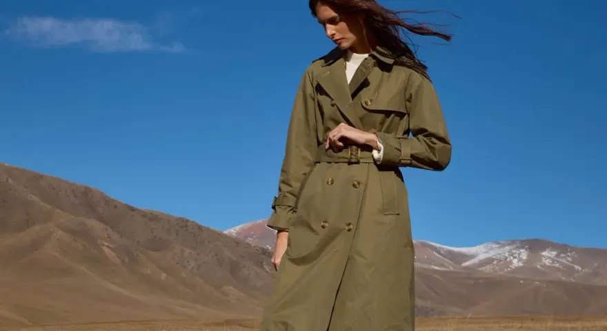 Mujer con un trench verde oliva, cinturón anudado, en un paisaje montañoso con cielo azul.
