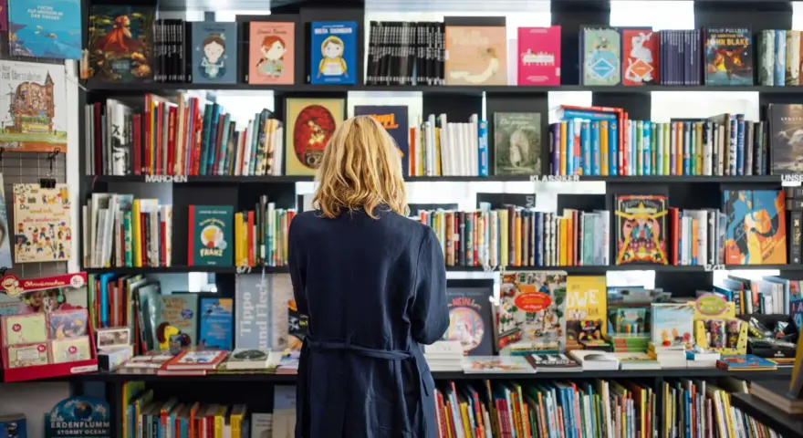 Donna bionda in libreria, circondata da scaffali pieni di libri colorati. Un'atmosfera di lavoro e scoperta tra le pagine.