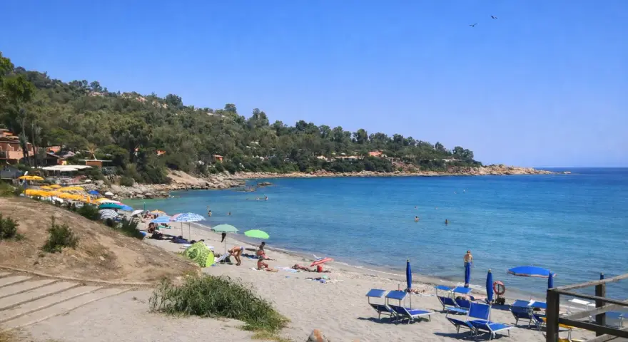 Spiaggia di Porto Frailis con ombrelloni colorati, lettini blu e gente che si gode il mare cristallino sotto il sole.