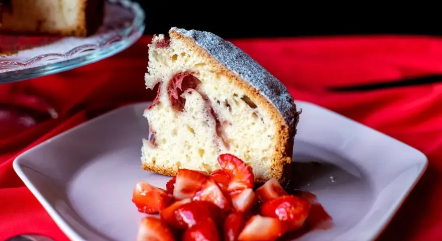 Un trozo de bizcocho de fresa esponjoso, espolvoreado con azúcar glas, servido con fresas frescas cortadas en un plato rosa.
