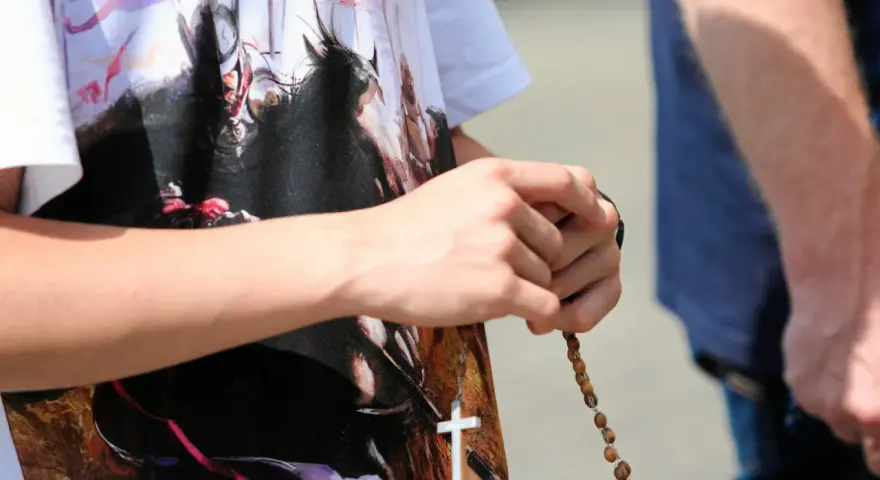 Dłoń trzyma paciorki różańca, a na koszulce widnieje biały krzyż. To znak modlitwy, żeby się wszystko ułożyło.