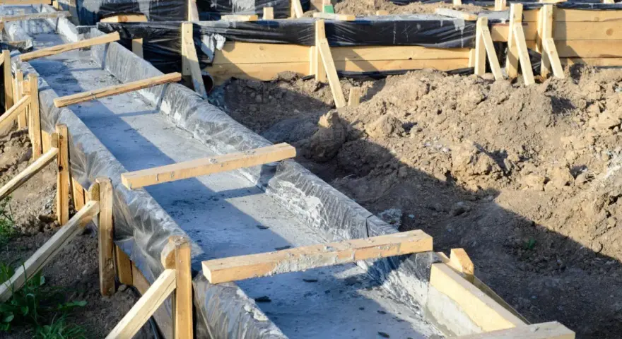 Fundamenty wylewane w wykopie, zabezpieczone folią, z drewnianym szalunkiem. Widać ziemię i przygotowania do dalszych prac budowlanych, uwzględniające głębokość przemarzania gruntu wzór.