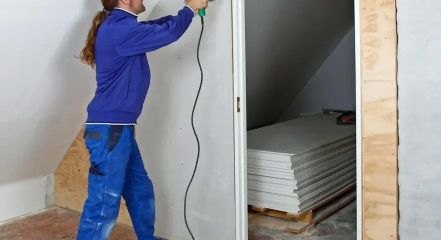 Man med skruvdragare monterar gipsskivor, kanske för att fixa en spackelremsa jula. Staplade skivor syns i bakgrunden.