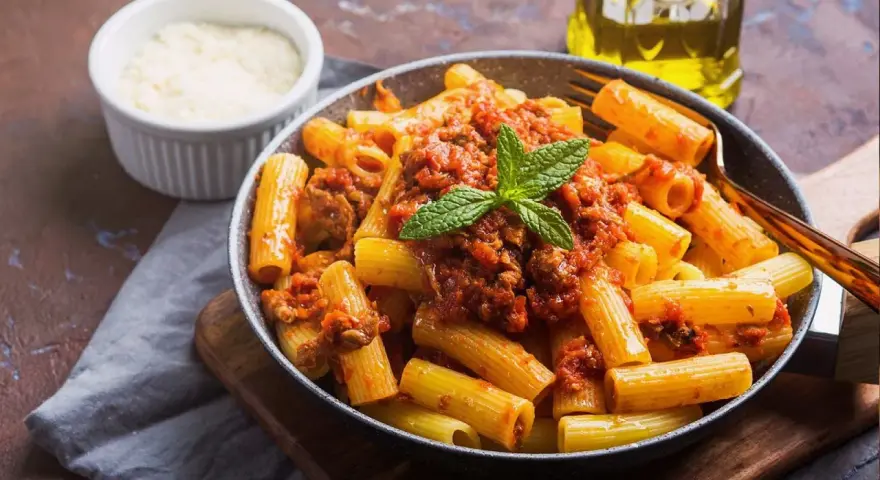 Un plato de macarrones con carne picada, cubiertos con salsa de tomate y decorados con menta. Al lado, un cuenco de queso rallado y una botella de aceite de oliva.