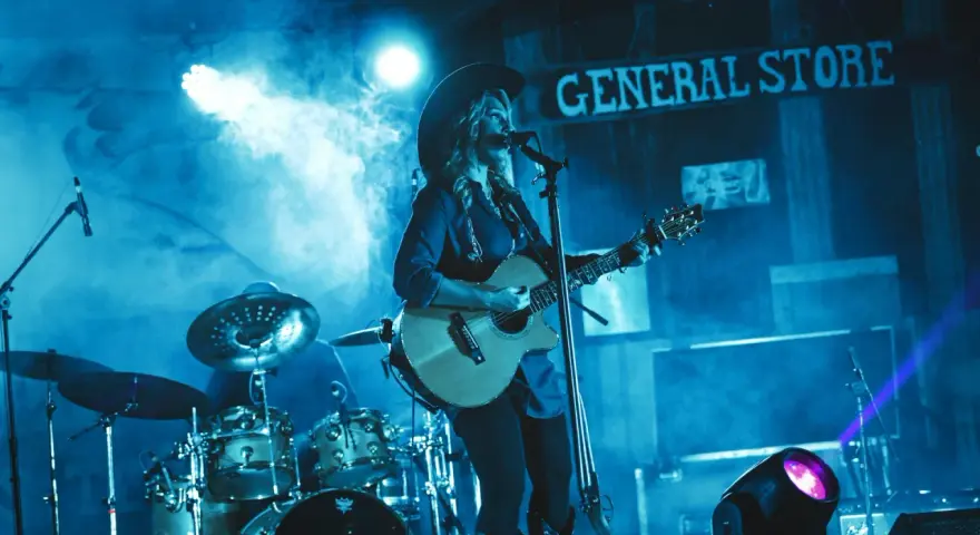 Artystka w kapeluszu gra na gitarze, wykonując najpopularniejsze piosenki country. W tle perkusja i napis "GENERAL STORE".