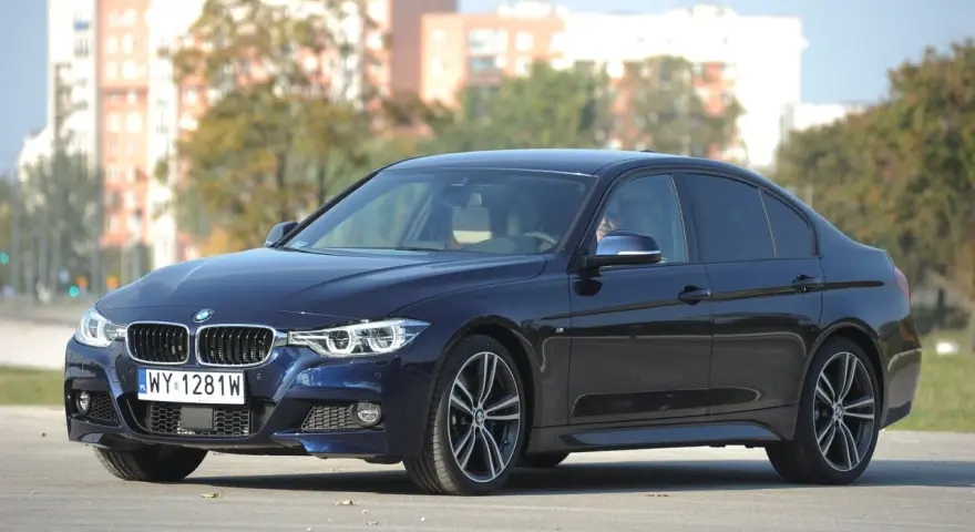 Ciemnoniebieskie BMW 318d z polskimi tablicami rejestracyjnymi stoi na tle budynków i drzew.