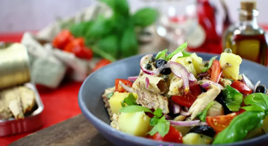Un'insalata pantesca colorata con patate, pomodorini, olive, cipolla rossa e pesce, guarnita con basilico fresco.