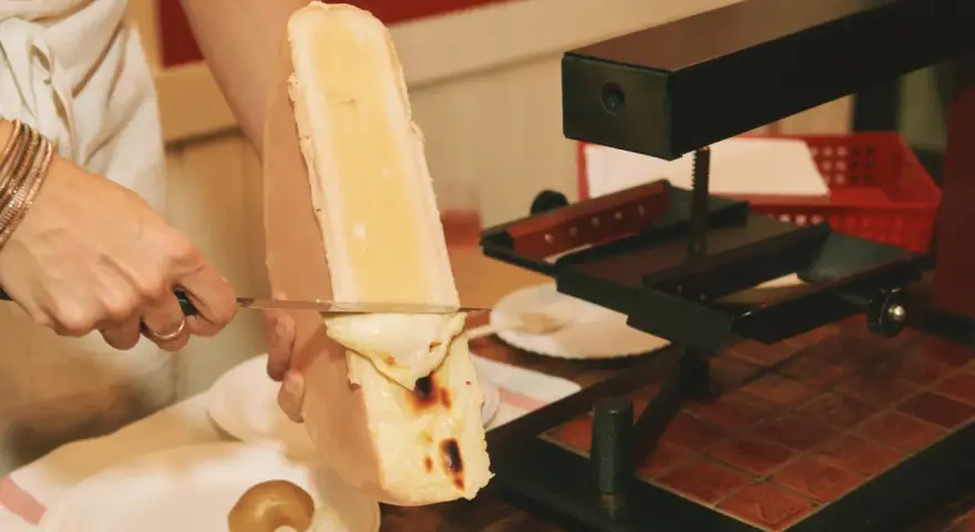 En person skrapar smält ost från en stor ostbit med en kniv. Perfekt för en mysig kväll med vin till raclette.