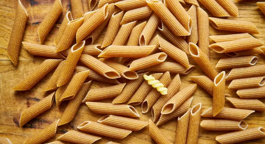 Makaron pełnoziarnisty vs zwykły. Na drewnianym tle leży mnóstwo makaronu penne. Wśród nich wyróżnia się jeden, żółty makaron fusilli.