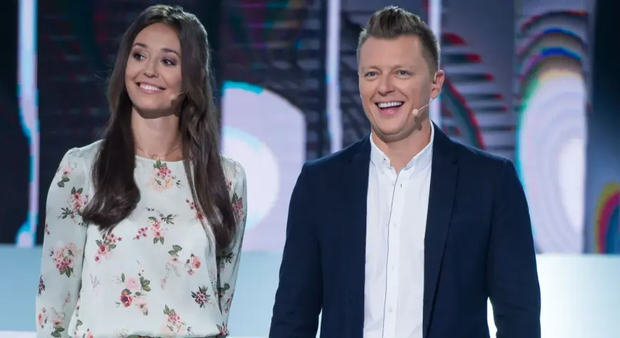 Uśmiechnięta para na scenie, gdzie nagrania programu telewizyjnego. Ona w kwiecistej sukience, on w marynarce.