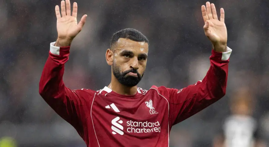 Mohamed Salah w czerwonej koszulce Liverpoolu, z uniesionymi dłońmi, pozdrawia kibiców. Widać logo klubu i sponsora.