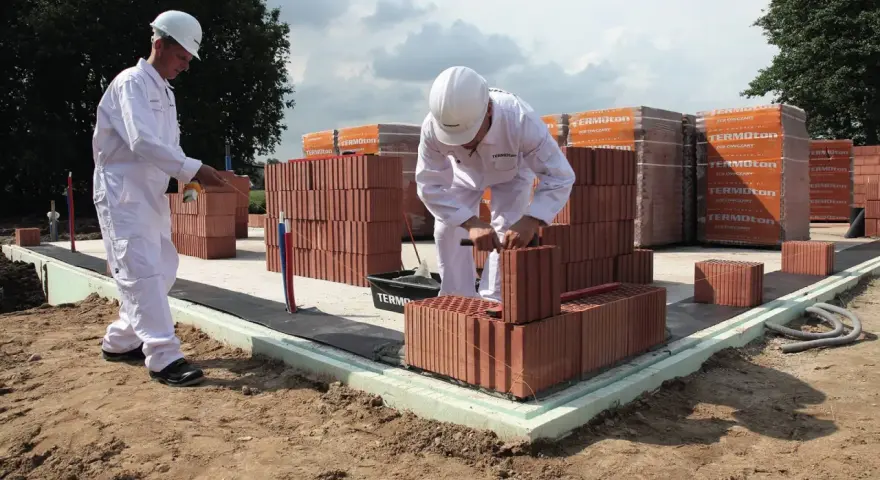 Dwóch budowlańców w białych kombinezonach układa cegły na fundamencie.