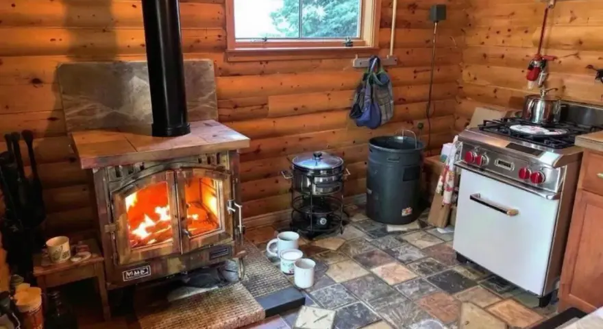 Poêle à bois allumé dans un chalet en bois rond, respectant la norme d'installation. Cuisine fonctionnelle avec cuisinière et ustensiles.