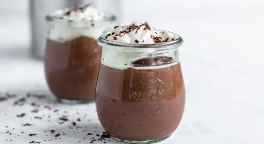 Zwei Gläser mit cremigem omas schokoladenpudding, gekrönt mit Schlagsahne und Schokostreuseln.