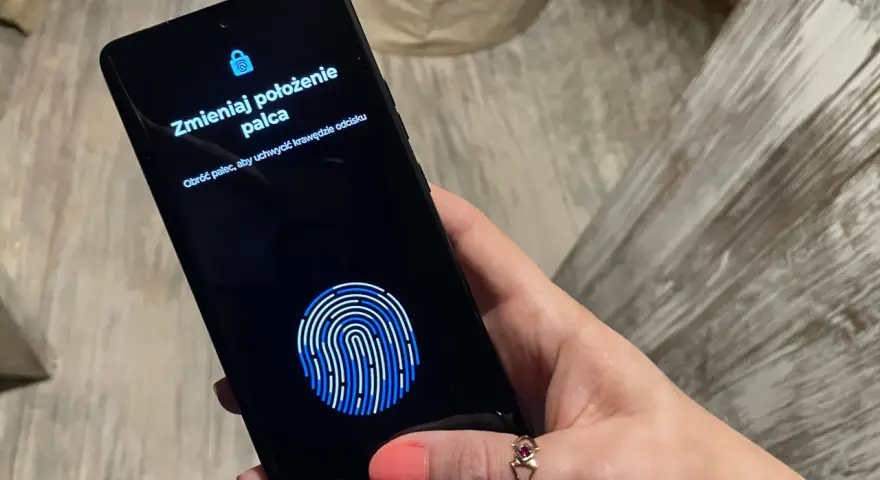 Dłoń z telefonem, na ekranie skaner linii papilarnych i komunikat "Zmieniaj położenie palca". To jak próba ucieczki z cyfrowego więzienia, niczym **jailbreak**.