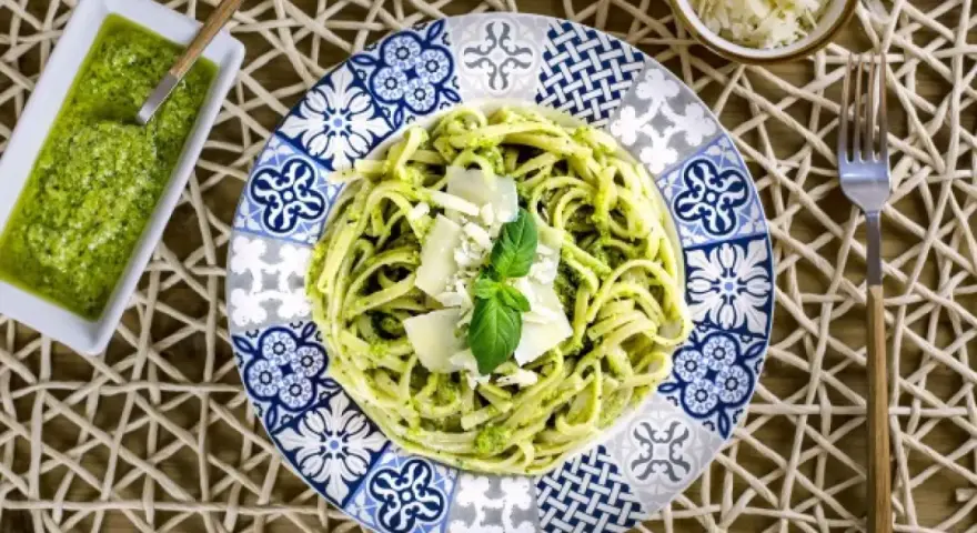 Pasta al pesto servida en plato decorado, con salsa de pesto y queso rallado al lado.