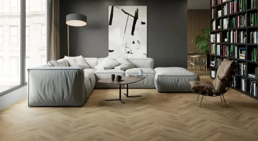 Nowoczesny salon z szarą kanapą i regałem pełnym książek. Podłoga w jodełkę imituje drewno, pokazując, jak dobrać płytki drewnopodobne do paneli.