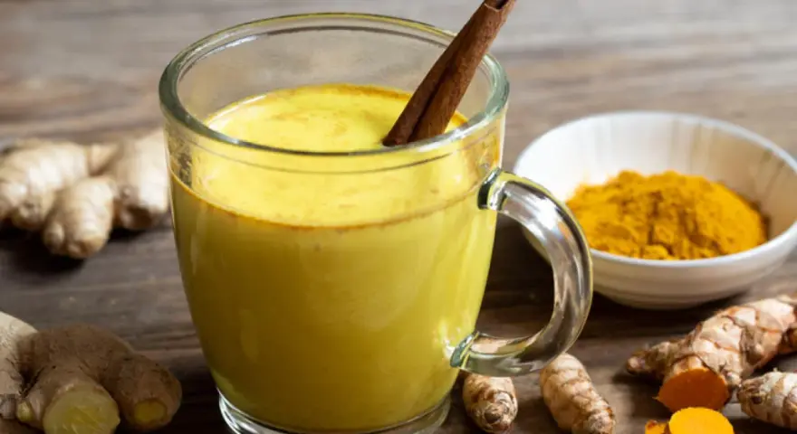 Taza de leche dorada con canela, rodeada de raíz de cúrcuma fresca y en polvo.