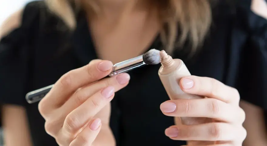 Dłonie z perfekcyjnym manicure nakładają pędzelkiem dr irena eris podkład rozświetlający na pociągłą tubkę.