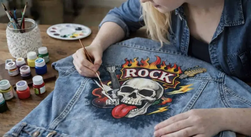 Dziewczyna maluje na dżinsowej kurtce czaszkę z płomieniami i napisem "ROCK". Czy farba akrylowa nadaje się do tkanin? Tak, jak widać na zdjęciu!