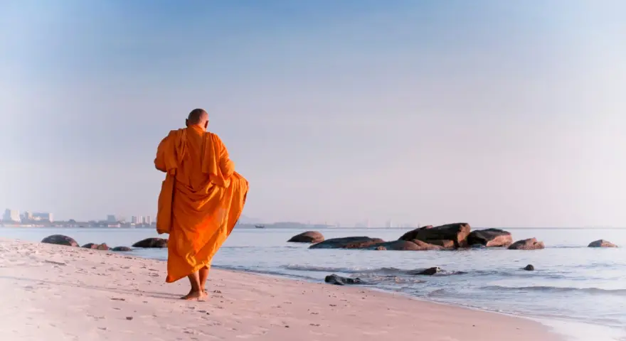 A modern day monk in an orange robe walks along a serene beach at sunrise, with calm ocean waves and distant city skyline.