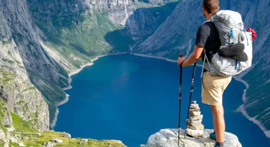 Un escursionista con zaino e bastoncini ammira un lago blu intenso circondato da montagne imponenti. Un'avventura di trekking montagna indimenticabile.