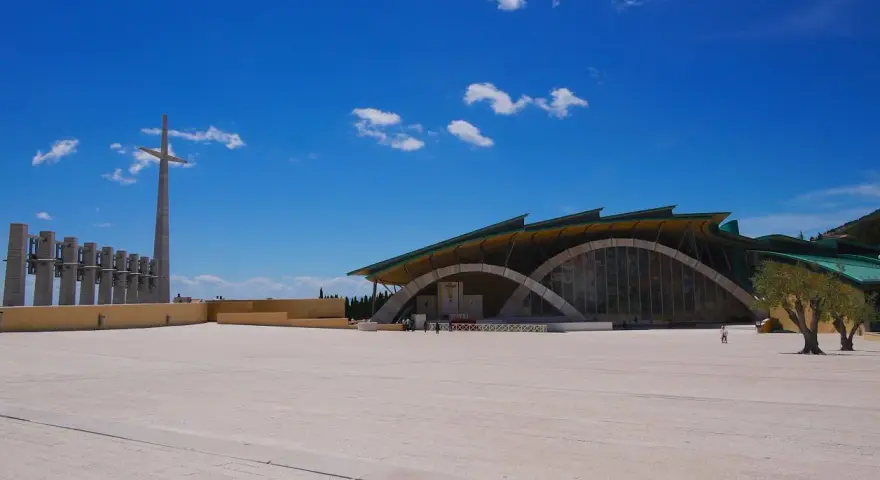 The modern church of San Giovanni Rotondo Padre Pio stands under a bright blue sky, with a tall cross and bell tower in the distance.